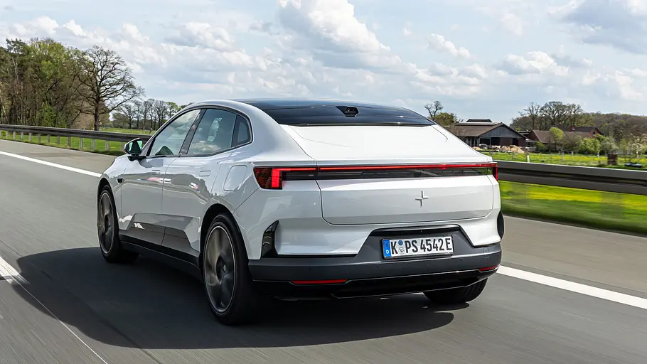 Ein weißer Polestar 4 fährt auf einer Straße mit Leitplanken, von schräg hinten gesehen. Im Hintergrund stehen Bäume und Häuser.