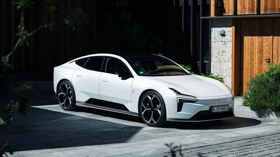Ein weißer Polestar 5 Performance mit schwarzem Glasdach steht auf einer gepflasterten Fläche vor einem Gebäude mit Holzfassade.