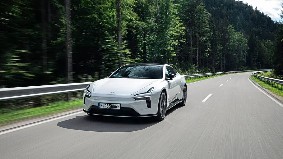 Ein weißer Polestar 5 Performance fährt auf einer kurvigen Straße mit Leitplanken durch eine dicht bewaldete Hügellandschaft.