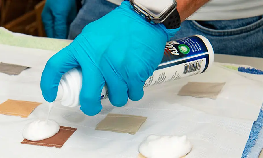 Eine Hand mit Gummihandschuh bekleidet sprüht einen Polsterreiniger auf quadratische Testflecken.