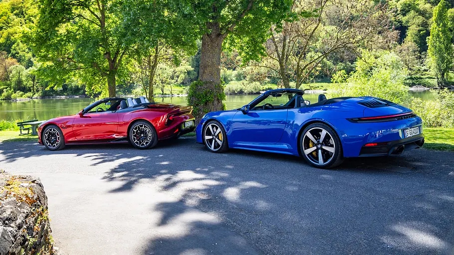 Das Porsche 911 Cabrio und der MG Cyberster stehen hintereinander am Rand einer Landstraße, im Hintergrund ein Fluss. Fotografiert von schräg hinten.