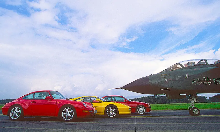 Der Porsche 911 Carrera 2/Ferrari F355/BMW M3 (E36) statisch von der Seite fotografiert. Ihnen gegenüber ein Tornado-Kampfjet.