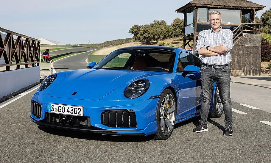 AUTO ZEITUNG-Redakteur Martin Urbanke steht auf Höhe der Fahrertür am Porsche 911 Turbo S (2025), fotografiert von schräg vorne.