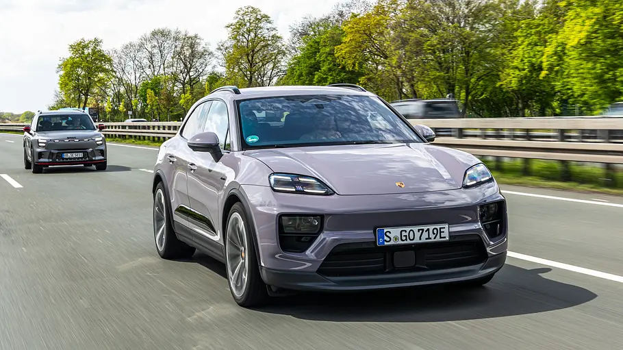 Ein hellvioletter Porsche Macan 4 fährt auf einer Autobahn. Dahinter folgt ein weiteres Auto, am Straßenrand stehen grüne Bäume.