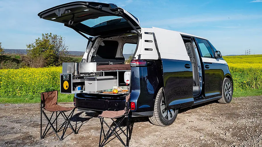 Eine Ququq Campingbox im Heck eines VW ID. Buzz Cargo. Das von schräg hinten rechts gezeigte Fahrzeug steht mit geöffneter Heckklappe vor einem Rapsfeld, zwei Campingstühle stehen am Heck.