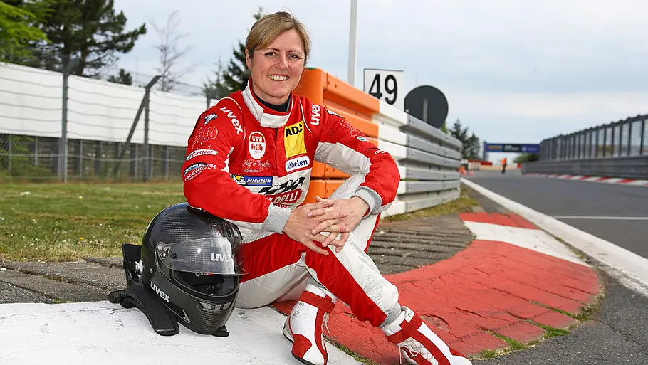 Blick von vorne auf Sabine Schmitz, die auf den Curbs der Nordschleife sitzt und in die Kamera lächelt.