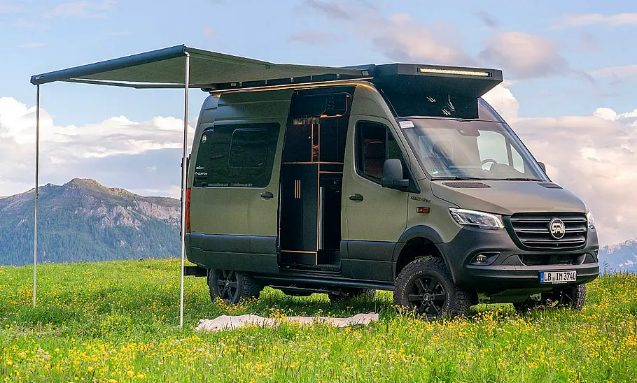Sam the Van (2025) Sam the Van (2025) auf einer Wiese stehend mit ausgefahrener Markise von schräg vorne rechts fotografiert.