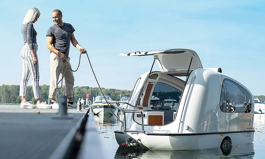 Sealander (2024) Der schwimmende Sealander (2024) an einem Bootssteg. Eine Person hält ihn an einem Seil fest. Eine zweite Person steht daneben.