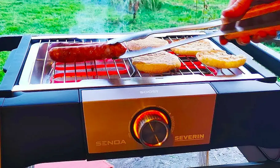 Severin Senoa Boost S Der Severin Senoa Boost S mit aufgelegten Speisen von vorne fotografiert. Eine Hand wendet mittels Grillzange eine Wurst.