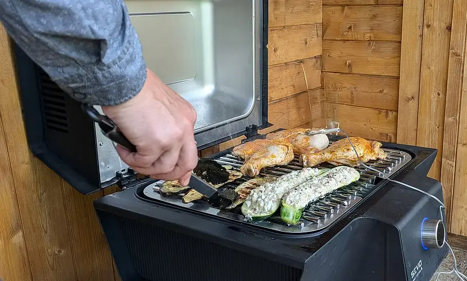 Severin Sevo Smart Control Der Severin Sevo Smart Control mit geöffnetem Deckel und aufgelegten Speisen von schräg vorne fotografiert. Eine Hand wendet das Grillgut mit einer Grillzange.