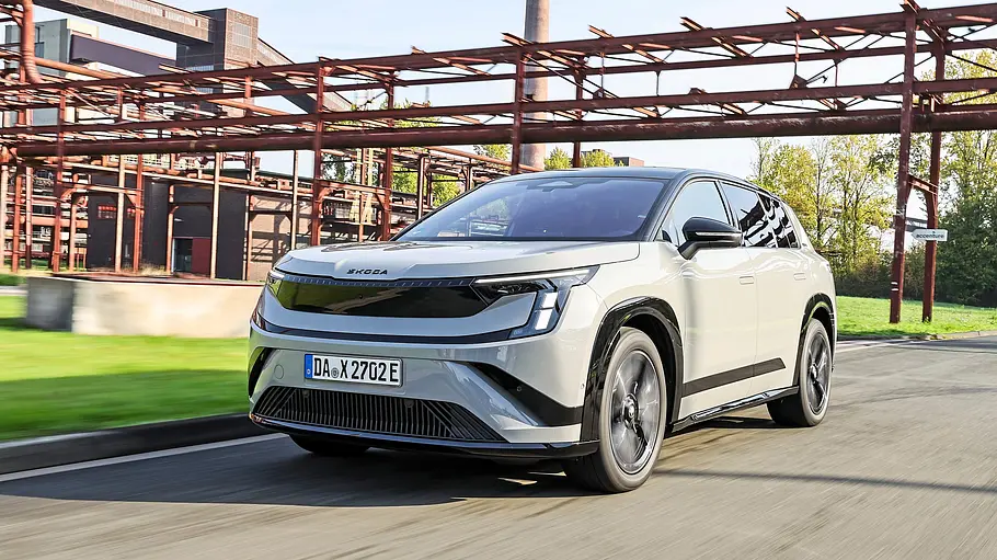 Hellgrauer Skoda Peaq fährt schräg von vorn unter rostigen Rohrbrücken eines Industriegeländes.