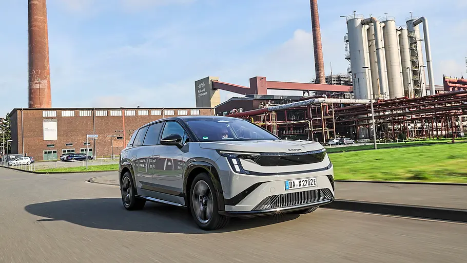 Skoda Peaq in Hellgrau fährt vor Backsteinhallen, Schornsteinen und Industrieanlagen schräg von vorn.