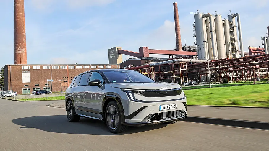 Skoda Peaq in Hellgrau fährt vor Backsteinhallen, Schornsteinen und Industrieanlagen schräg von vorn.