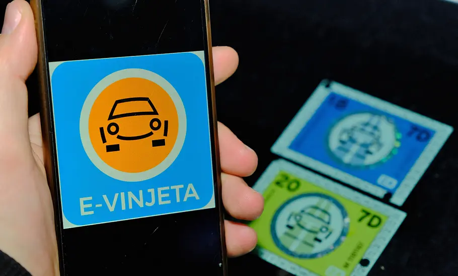 E-Vinjeta für Slowenien Eine Hand hält ein Smartphone auf dem ein Auto-Symbol und E-Vinjeta zu sehen ist. Unscharf im Hintergrund liegen alte Klebevignetten.