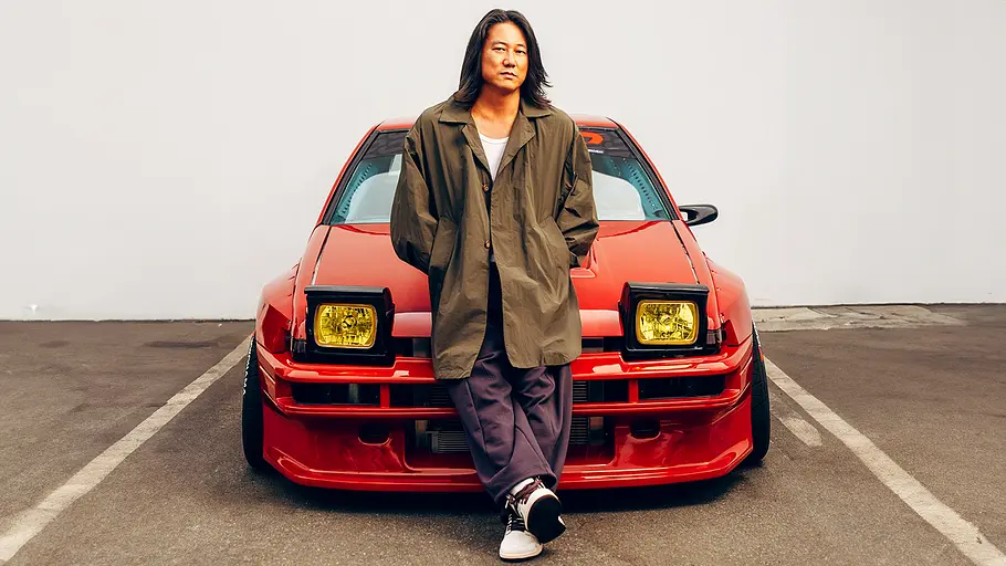 Sung Kang, der sich an die Motorhaube eines Toyota AE86 lehnt, von vorne fotografiert.