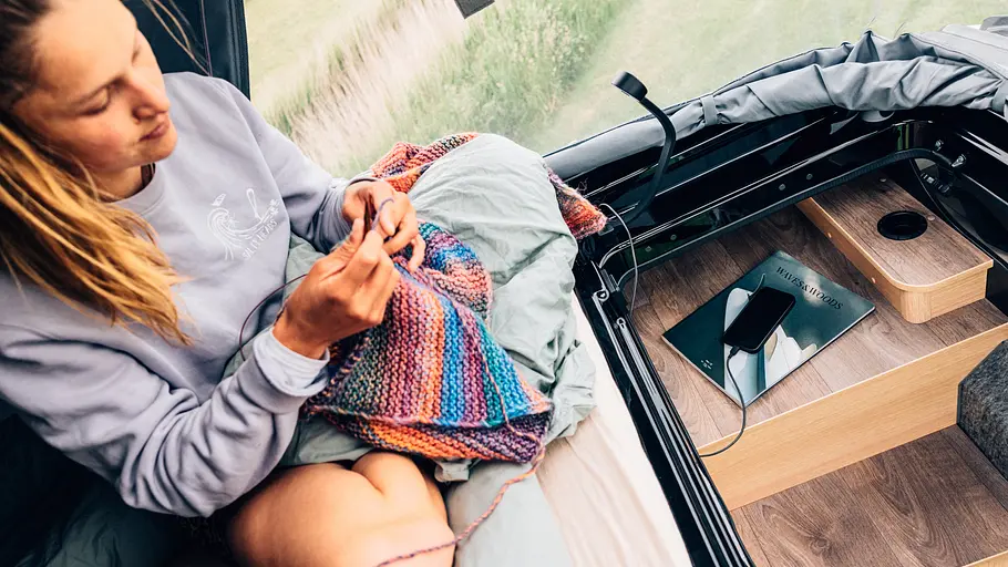 Sunlight Cliff 540V Vanlife Eine Person häckelt sitzend auf der Matzratze im Schlafdach. Auf der obersten Stufe liegt ein Magazin und ein Smartphone mit Ladekabel.