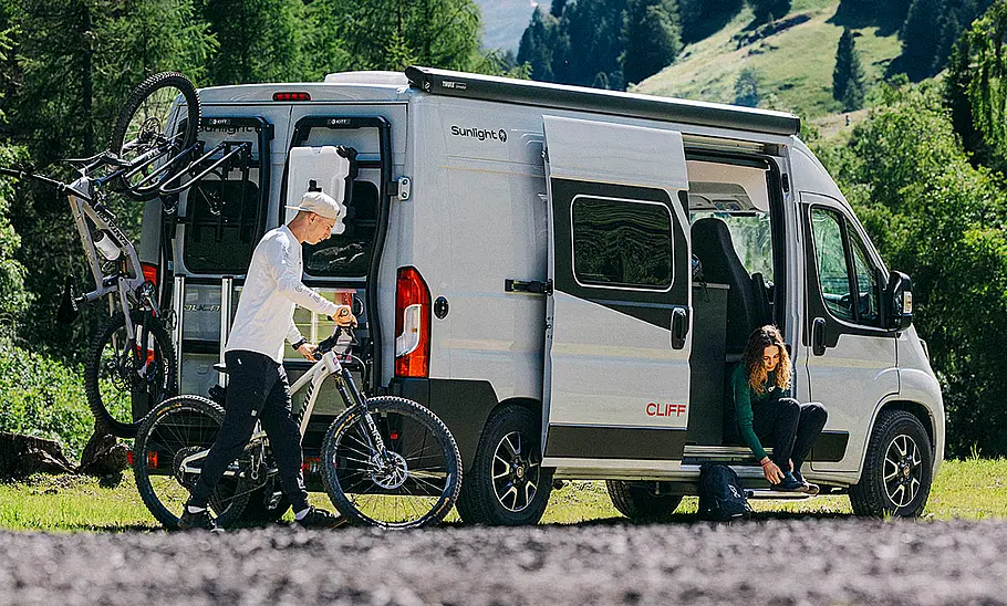 Das Bild zeigt den Sunlight Cliff Adventure 602 stehend von schräg hinten. Eine Person nimmt ein Fahrrad vom Fahrradträger, eine zweite sitzt im Bereich der geöffneten Schiebetüre.