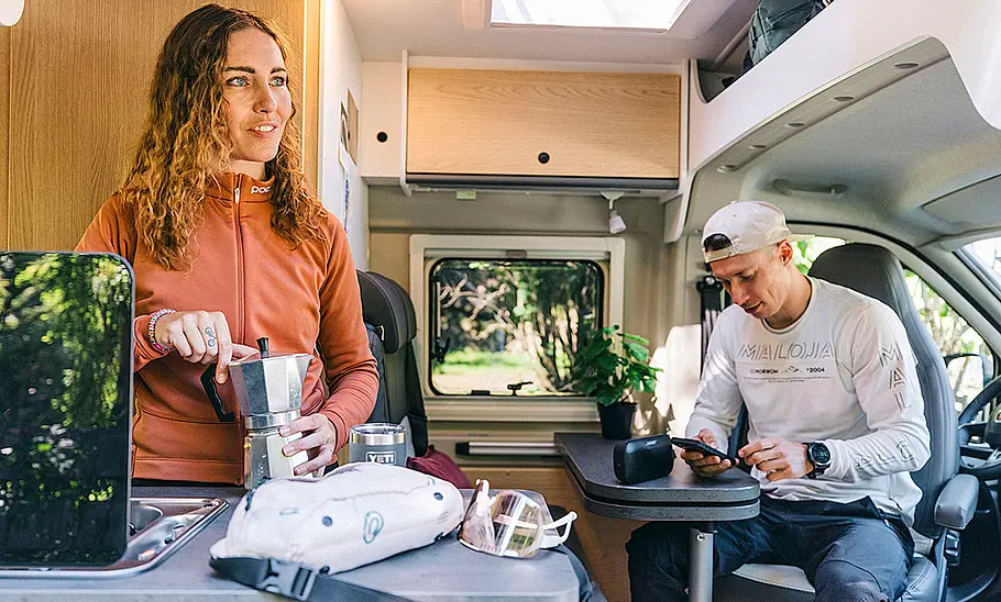 Das Bild zeigt den Wohnraum des Sunlight Cliff Adventure 602 von der geöffneten Schiebetüre aus. Eine am Herd stehende Person schraubt einen Espressokocher zusammen, eine andere Person sitzt an der Sitzgruppe.