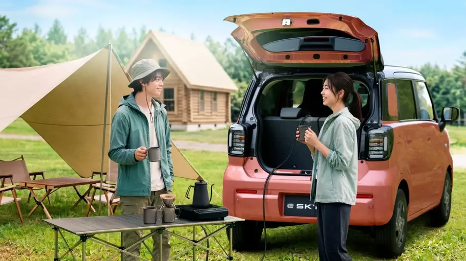 Suzuki e Sky Zwei Personen mit Bechern stehen neben dem Suzuki e Sky mit offener Heckklappe. Davor steht ein Campingtisch mit Kaffeekochzubehör.