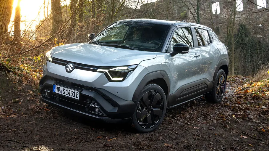 Der Suzuki e Vitara steht in einem Waldstück, von vorne links fotografiert.