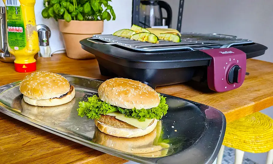 Tefal Easygrill Der Tefal Easygrill von schräg vorne fotografiert im Hintergrund. Vorne ein Tablett mit zwei Burgern.