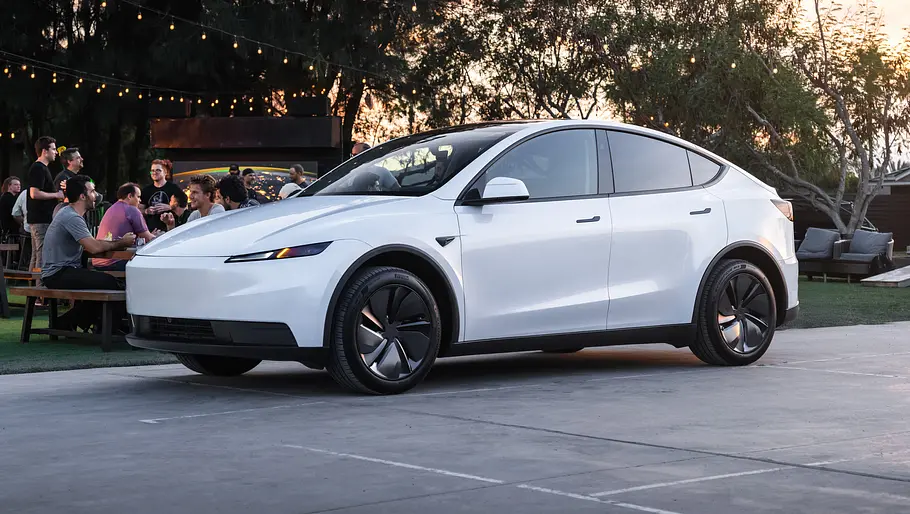 Tesla Model Y Facelift Das Tesla Model Y Facelift steht auf einem Parkplatz, im Hintergrund feiern Menschen eine Gartenparty.