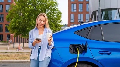 Eine Frau steht mit einem Coffee-To-Go-Becher und einem Smartphone in der Hand neben einem Skoda Enyaq Coupé, das per Kabel am Straßenrand aufgeladen wird. - Foto: Imago