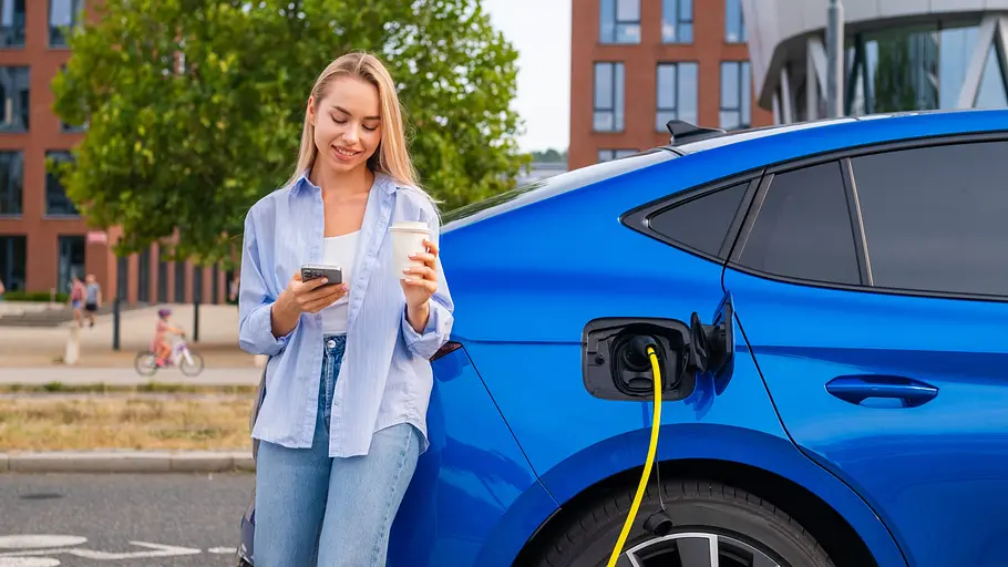Tipps und Tricks für E-Auto-Neulinge Eine Frau steht mit einem Coffee-To-Go-Becher und einem Smartphone in der Hand neben einem Skoda Enyaq Coupé, das per Kabel am Straßenrand aufgeladen wird.