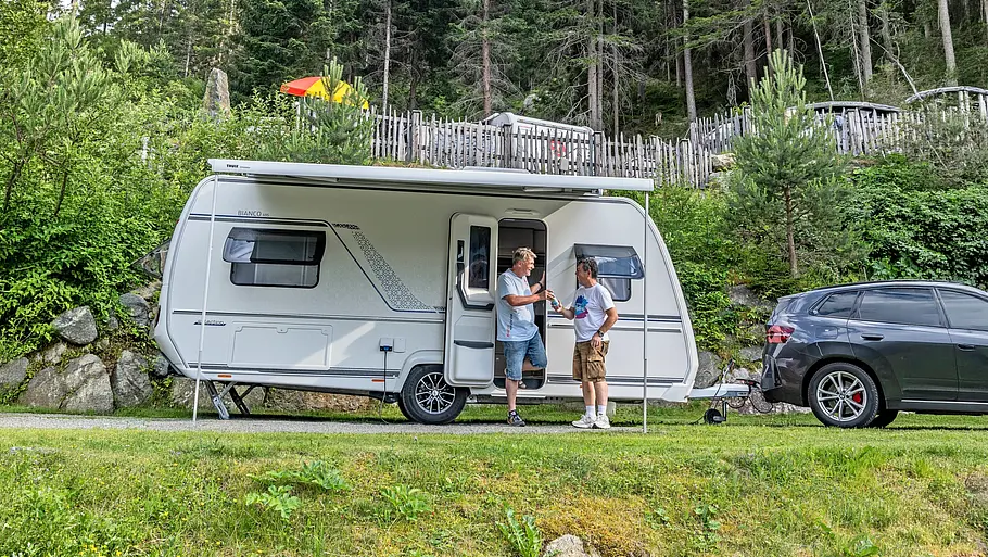 Tirol-Reisereportage im Fendt Bianco Das Gespann aus BMW X3 und Fendt Bianco auf dem Naturcampingplatz Kuprian von der Seite. Unter der Markise des Wohnwagens unterhalten sich zwei Personen vor der geöffneten Türe.