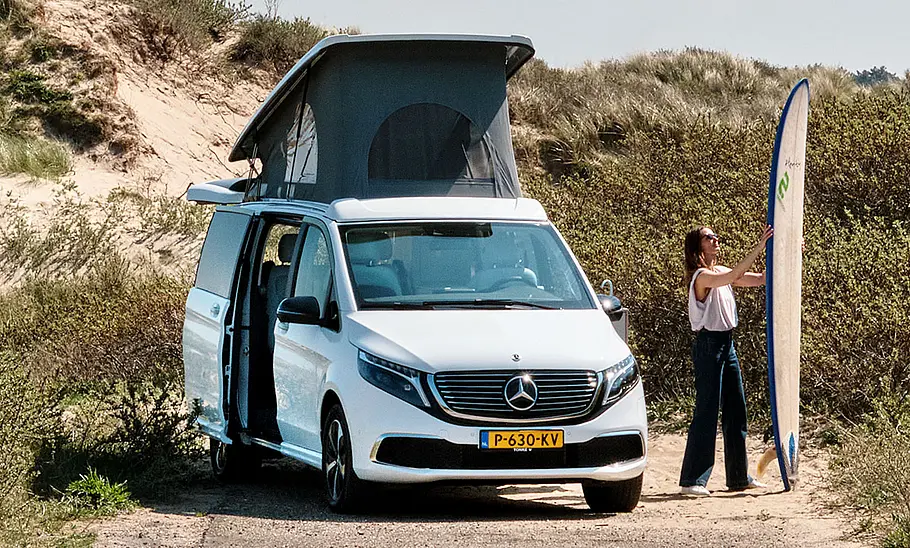 Das Bild zeigt den Camper in den Dünen stehend mit geöffnetem Aufstelldach. Daneben steht eine Person mit einem Surfbrett.