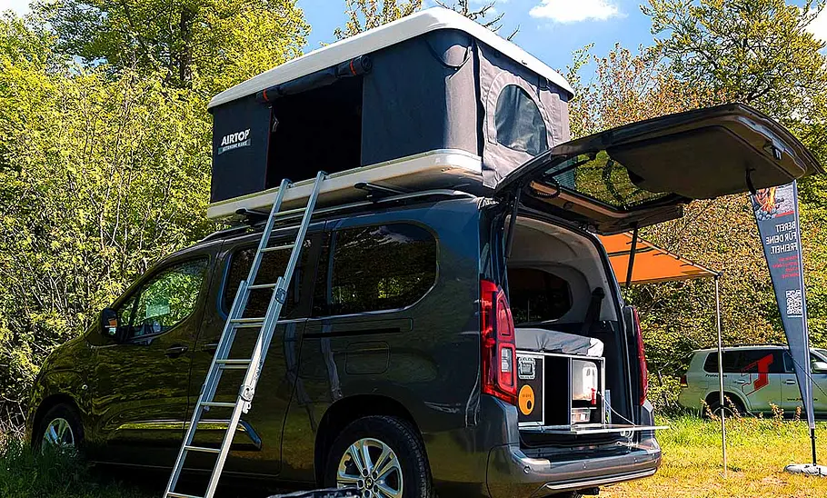 Toyota-Campingzubehör; Toyota Proace City Verso, stehend, mit offener Heckklappe. Im Van befindet sich eine Campingbox, auf dem Dach ist ein Dachzelt angebracht, eine Leiter ist an das Auto gelehnt.