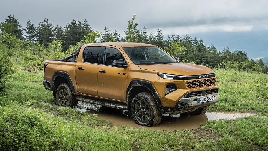 Toyota Hilux 2.8 D Der Toyota Hilux 2.8 D statisch von schräg vorne rechts mit Matschspritzern in einer Pfütze auf einer Wiese.