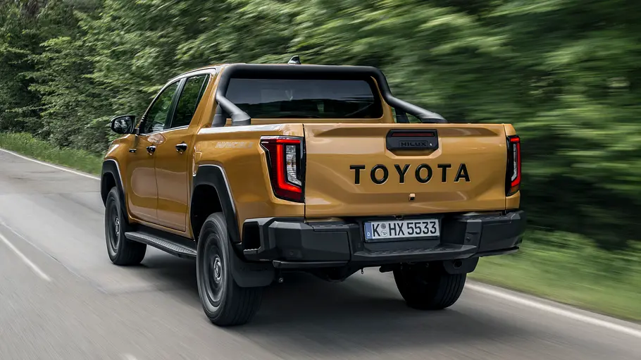 Toyota Hilux 2.8 D Der Toyota Hilux 2.8 D fahrend auf einer Landstraße durch ein Waldstück von schräg hinten links.