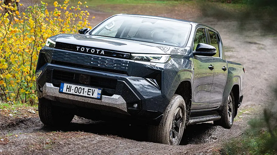 Der Toyota Hilux BEV fährt im Schlamm einen unebenen Hang hinauf, von vorne links fotografiert.