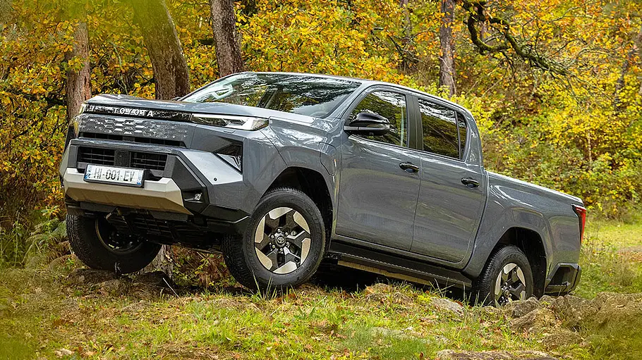 Der Toyota Hilux BEV steht an einem Wald, von vorne links fotografiert.