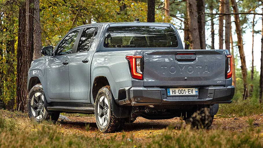 Der Toyota Hilux BEV steht in einem Wald, von hinten links fotografiert.