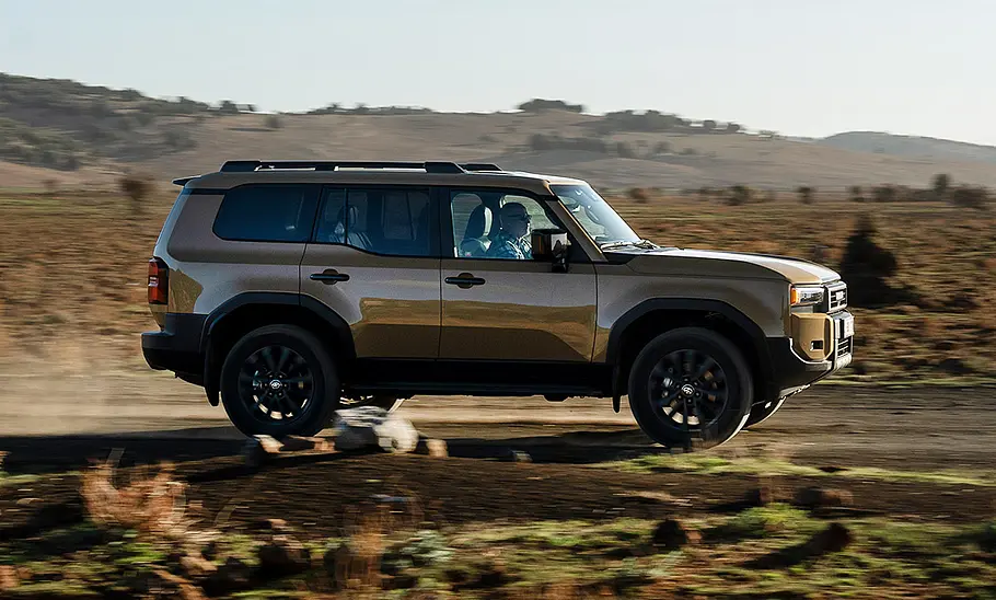 Toyota Land Cruiser (2024) Der Toyota Land Cruiser (2024) fährt über eine staubige Piste; von der Seite fotografiert.