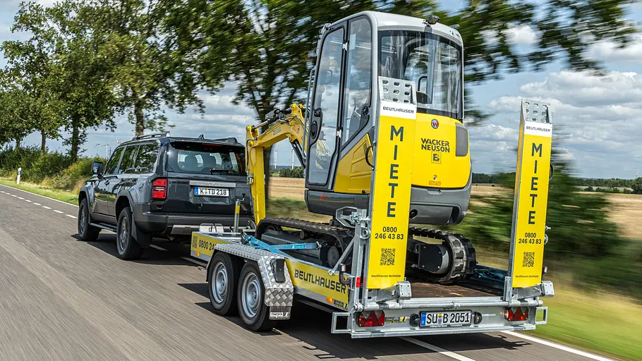 Toyota Land Cruiser Der Toyota Land Cruiser zieht einen Anhänger mit einem Bagger darauf, fotografiert von schräg hinten.