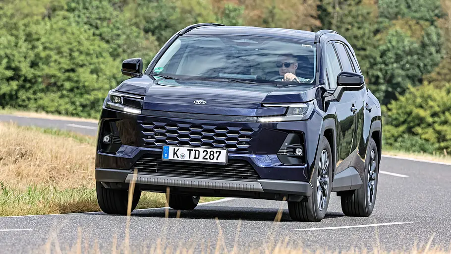 Toyota RAV4 und VW Tiguan im Vergleich Ein dunkelblauer Toyota RAV4 fährt auf einer kurvigen Landstraße, umgeben von trockenem Gras und grünen Bäumen.