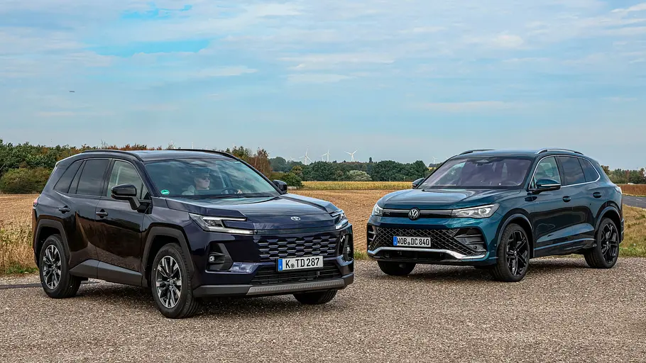 Toyota RAV4 und VW Tiguan im Vergleich Toyota RAV4 und VW Tiguan stehen nebeneinander auf einer Asphaltfläche vor Feldern unter einem weitläufigen, wolkigen blauen Himmel.