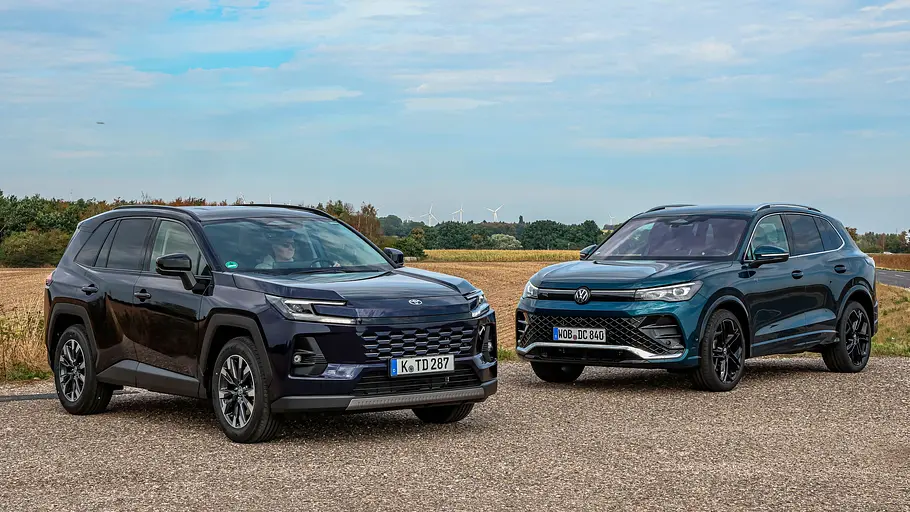 Toyota RAV4 und VW Tiguan Vergleichstest Toyota RAV4 und VW Tiguan stehen auf einer befestigten Fläche vor Feldern unter einem blauen Himmel mit weißen Wolken.