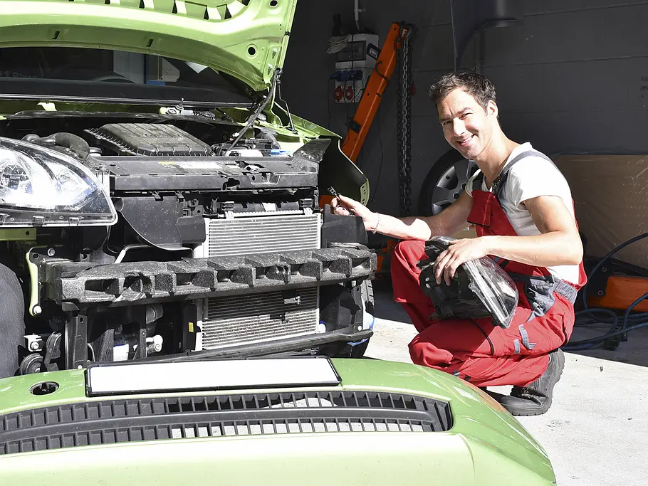 Eine Person arbeitet an einem Auto. Die Front ist demontiert. Die Person hat einen Scheinwerfer in der Hand.