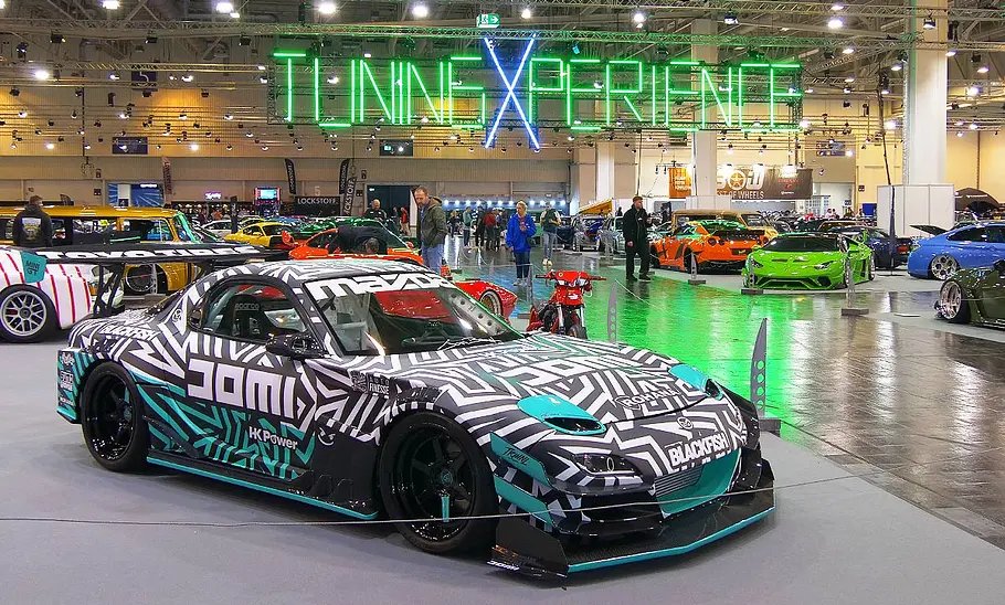 Blick von schräg vorne auf einen getunten Mazda RX-7. Im Hintergrund erstrecken sich die mit Autos gefüllten Hallen der Highlights der Tuning Xperience 2025.