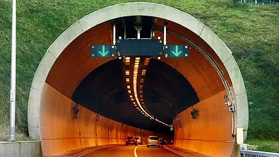 Eine Tunneleinfahrt in einem mit Gras bewachsenem Berg, die Röhre erscheint orangefarben beleuchtet. Über beiden Fahrspuren hängt eine Anzeige mit einem grünen Pfleit darauf, der nach unten zeigt. - Foto: Bauerstock/AUTO ZEITUNG
