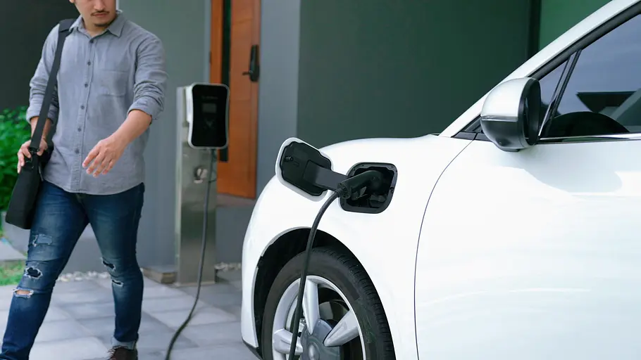 Ein Ausschnitt eines Elektroautos, das mit einer Wallbox verbunden ist. Daneben geht ein Mann vorbei.