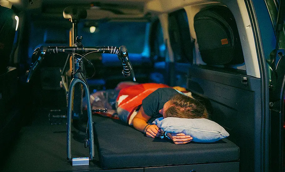 Das Bild zeigt den Bettumbau des Wohnmoduls des Vanderer Two (2024). Auf dem Bett liegt eine Person, daneben steht ein Fahrrad mit demontiertem Vorderrad im Innenraum des Fahrzeugs. Das Bild ist durch die geöffnete Kofferraumklappe aufgenommen worden.