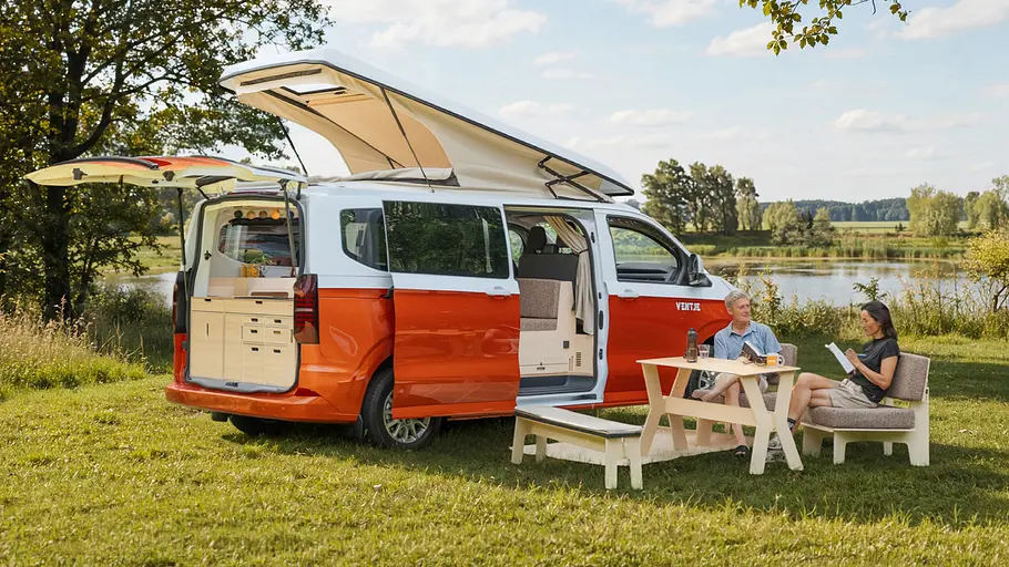 Der Ventje T7 Camper statisch von schräg hinten rechts, vor dem zwei Personen an einem Campingtisch sitzen.