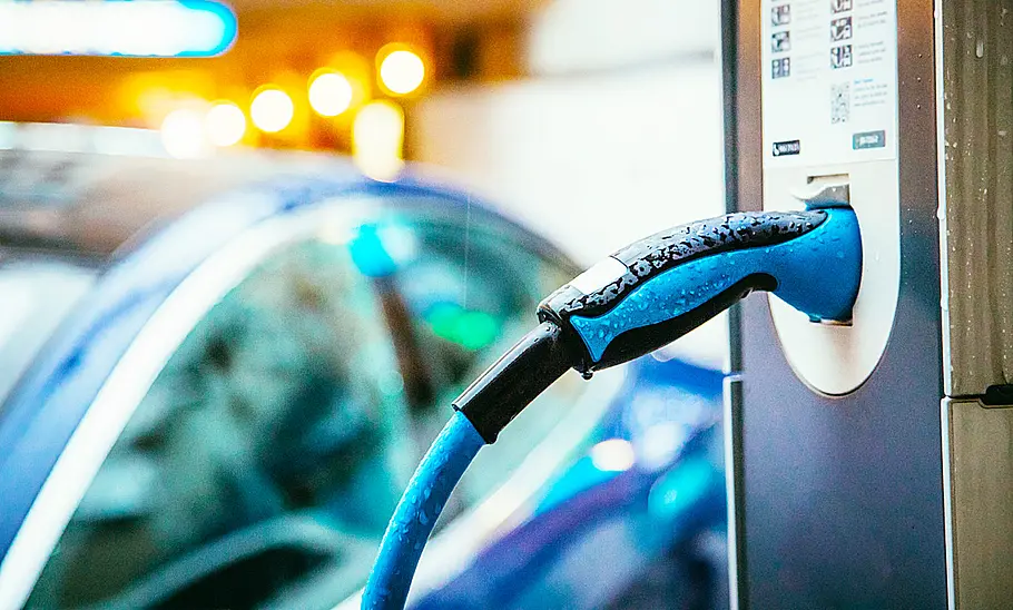 Ladestation mit angeschlossenem Kabel, im Hintergrund sieht man einen Ausschnitt von einem E-Auto.