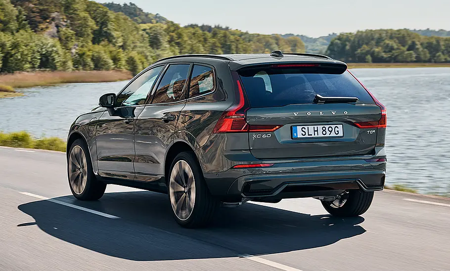 Der Volvo XC60 (2025) fährt auf einer Landstraße vor einem See, von schräg hinten fotografiert.