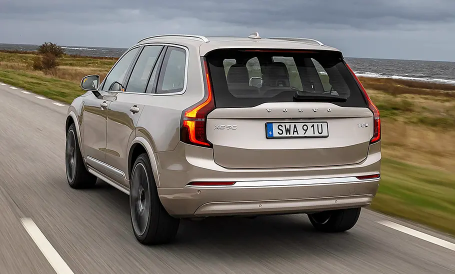 Das Volvo XC90 Facelift (2024) fährt über eine Landstraße, von schräg hinten fotografiert.